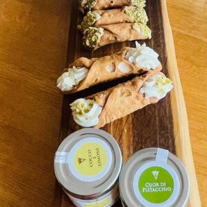 Cannoli fyldt med ricottacreme og pistacie ligger på et træfad ved siden af to glas med creme