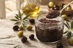 Åbent glas med mørk oliventapenade og en ske der løfter en portion op, med hele grønne og sorte oliven omkring